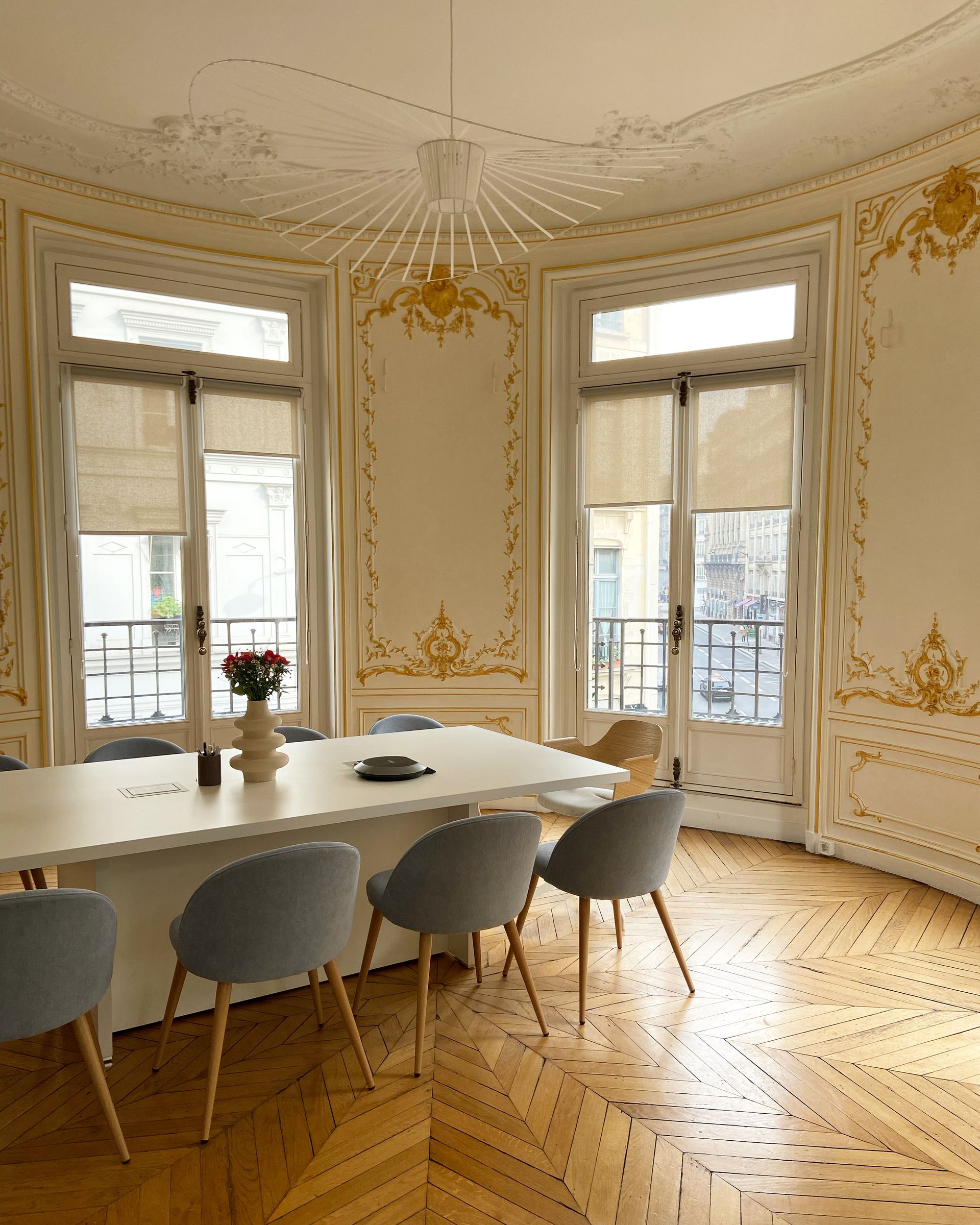 Une table à manger moderne avec des chaises grises, dans une pièce élégante et ornée de moulures, aux murs décorés de détails dorés et agrémentée de deux grandes fenêtres.