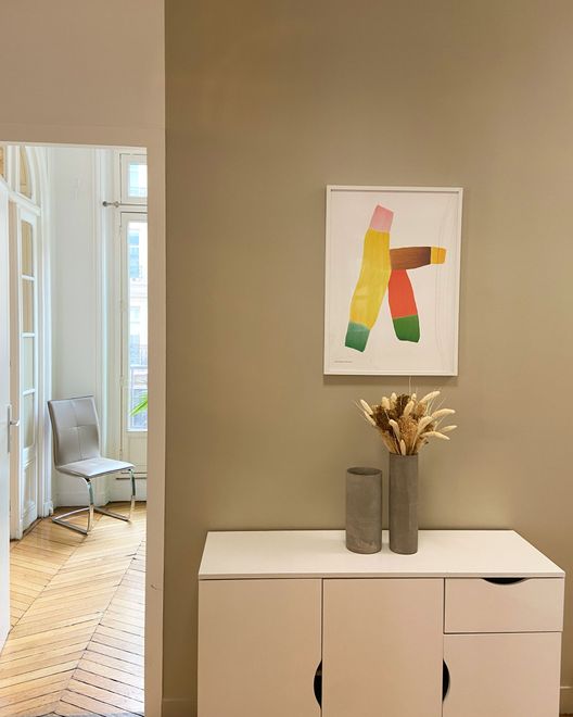 Un meuble blanc surmonté de deux vases contenant des fleurs séchées est placé sous une reproduction d'art abstrait colorée encadrée, accrochée au mur.