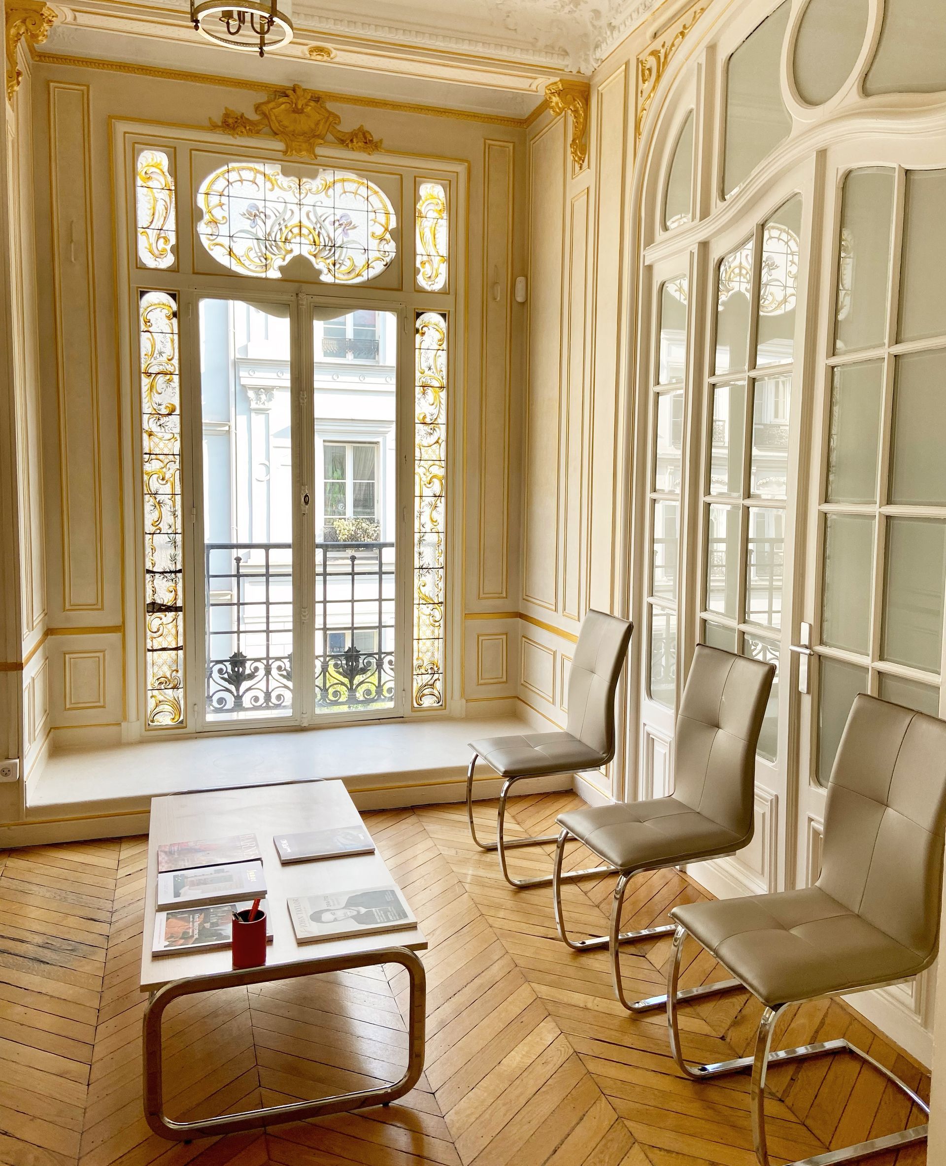 Salle d'attente avec trois chaises modernes grises, une table basse avec des magazines et de hautes portes-fenêtres à panneaux de verre finement ouvragées.