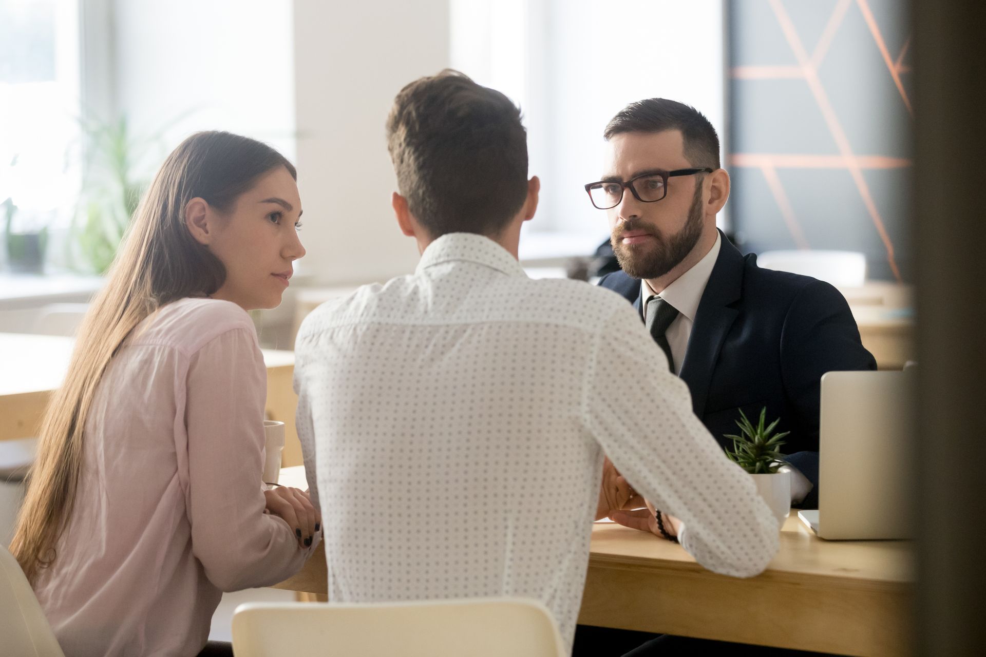 Un professionnel en costume discute avec deux personnes assises de part et d'autre d'une table en bois, dans un bureau.