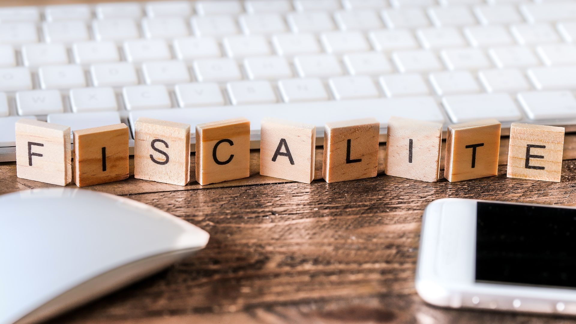 Des carreaux de bois formant le mot FISCALITÉ sont disposés sur un bureau en bois, avec un clavier d'ordinateur, une souris et un smartphone à proximité.