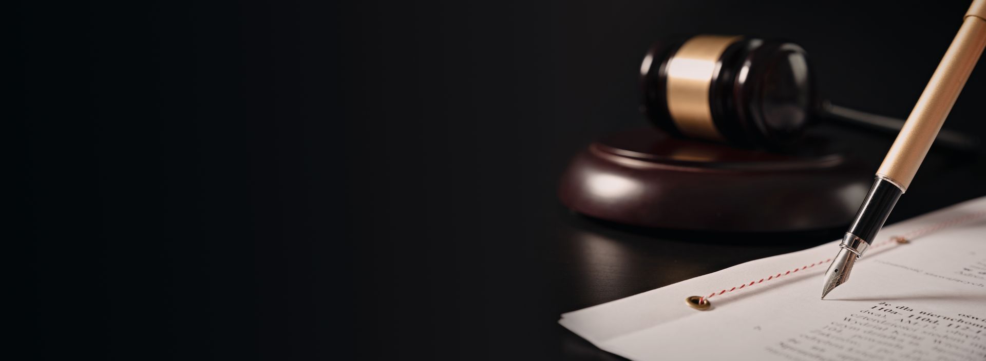 Un maillet repose sur un socle en bois à côté d'un stylo-plume posé sur un document juridique, le tout sur un fond sombre et minimaliste.
