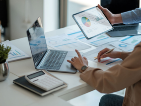 Une personne en costume pointe un stylo vers un document tenu par une autre personne en costume, assise à un bureau où se trouve une balance de la justice.