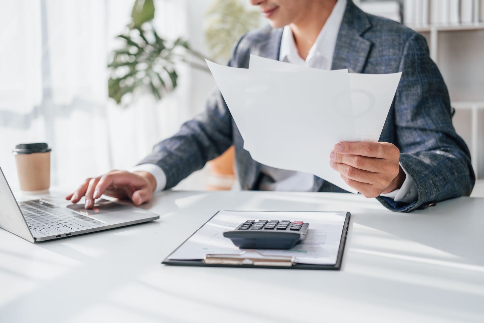 Une personne en costume, en train de consulter des documents à un bureau, avec un ordinateur portable et une calculatrice.