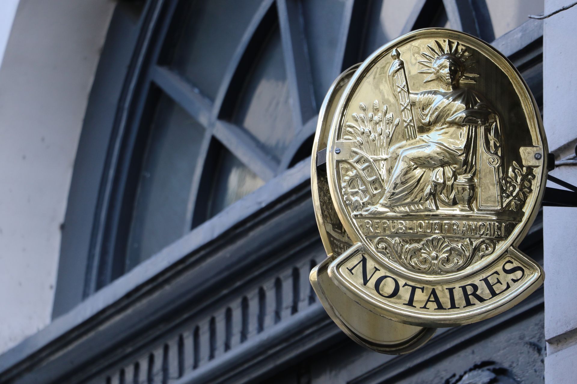 Une enseigne ovale dorée et brillante, ornée d'un emblème en relief, est accrochée au-dessus d'un encadrement de porte gris foncé. On peut y lire NOTAIRES.
