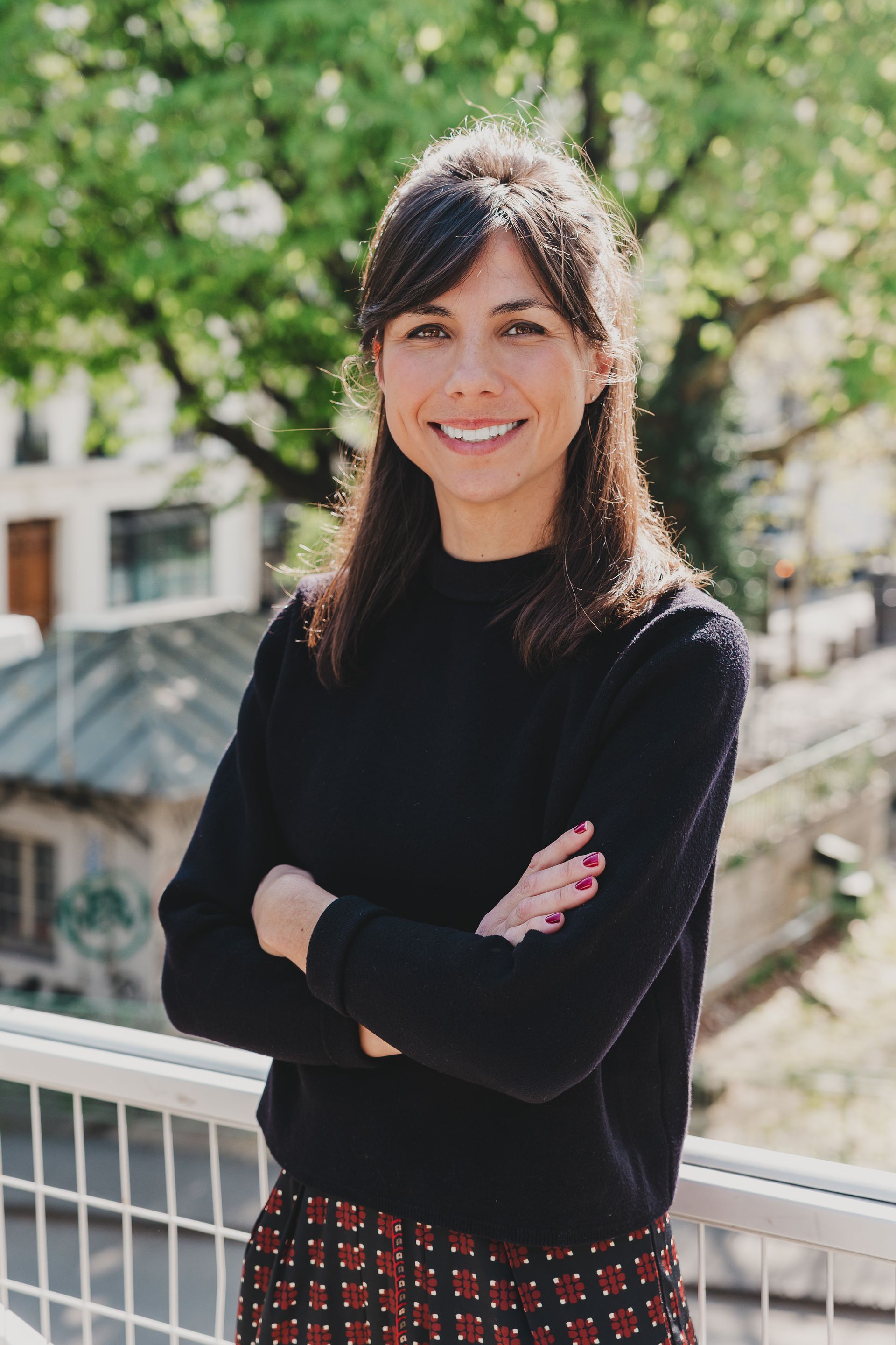 Une personne souriante, les bras croisés, vêtue d'un pull foncé et d'une jupe à motifs, se tient sur un balcon avec des arbres en arrière-plan.