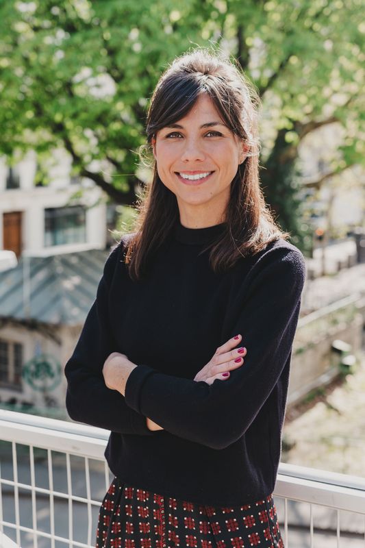 Une personne souriante, les bras croisés, vêtue d'un pull foncé et d'une jupe à motifs, se tient sur un balcon avec des arbres en arrière-plan.