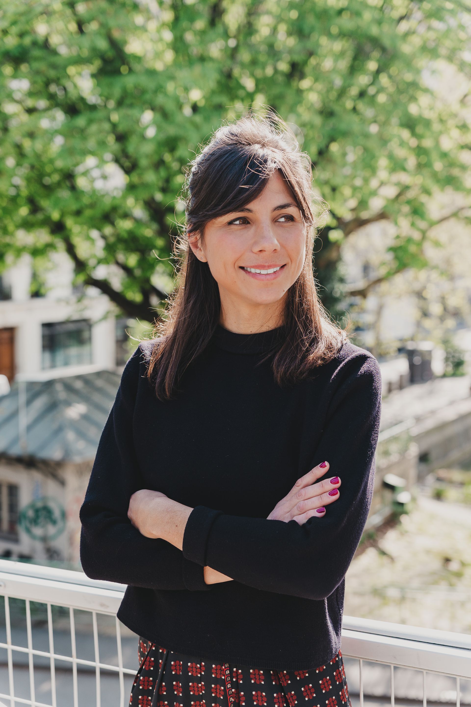 Une personne souriante aux longs cheveux noirs se tient debout, les bras croisés, devant un arrière-plan extérieur flou composé d'arbres et de bâtiments.