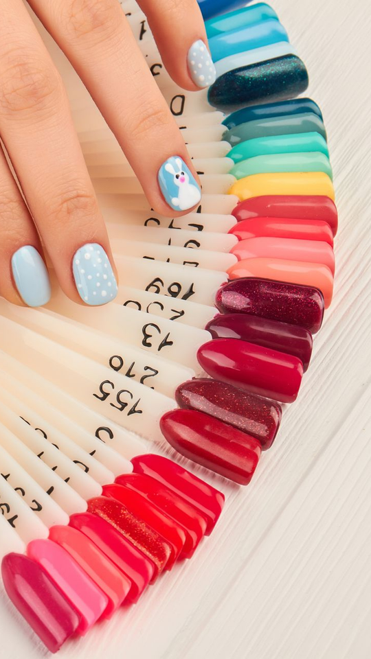 Une main ornée d'un nail art bleu clair et d'un lapin repose sur un éventail de nuanciers de vernis à ongles.