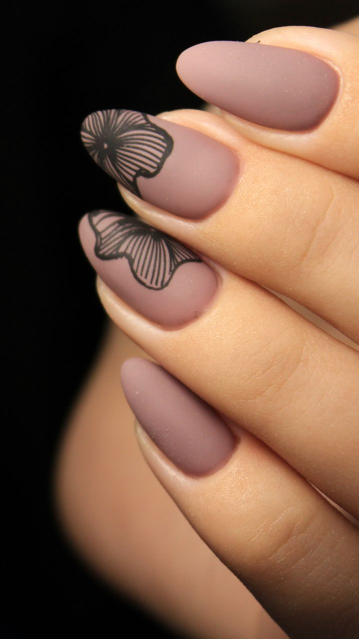Gros plan sur une main aux ongles en amande peints en mauve mat avec un motif floral noir sur deux ongles.