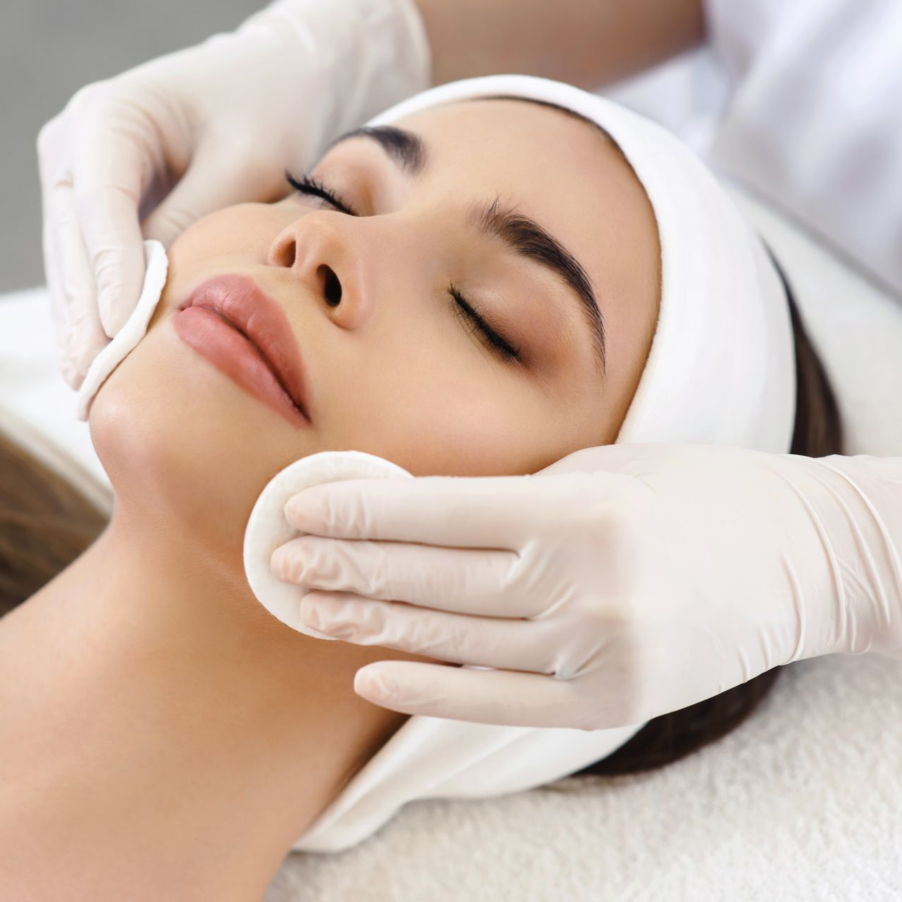 Femme recevant un soin du visage, mains gantées de blanc, yeux fermés, bandeau blanc, coussinets de coton.