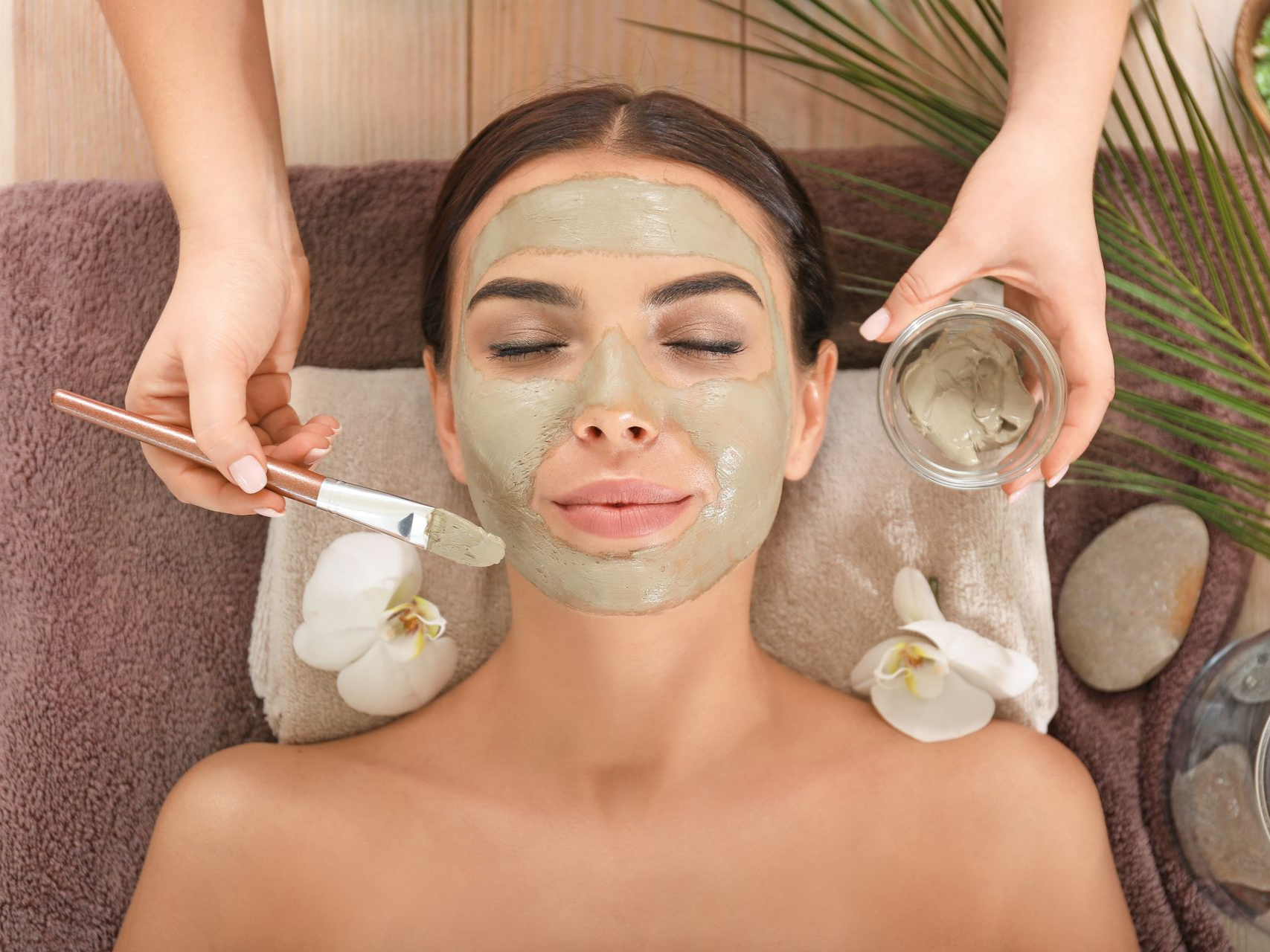 Une femme reçoit un masque facial dans un spa. Ses mains appliquent le masque au pinceau ; des orchidées et des pierres sont présentes.