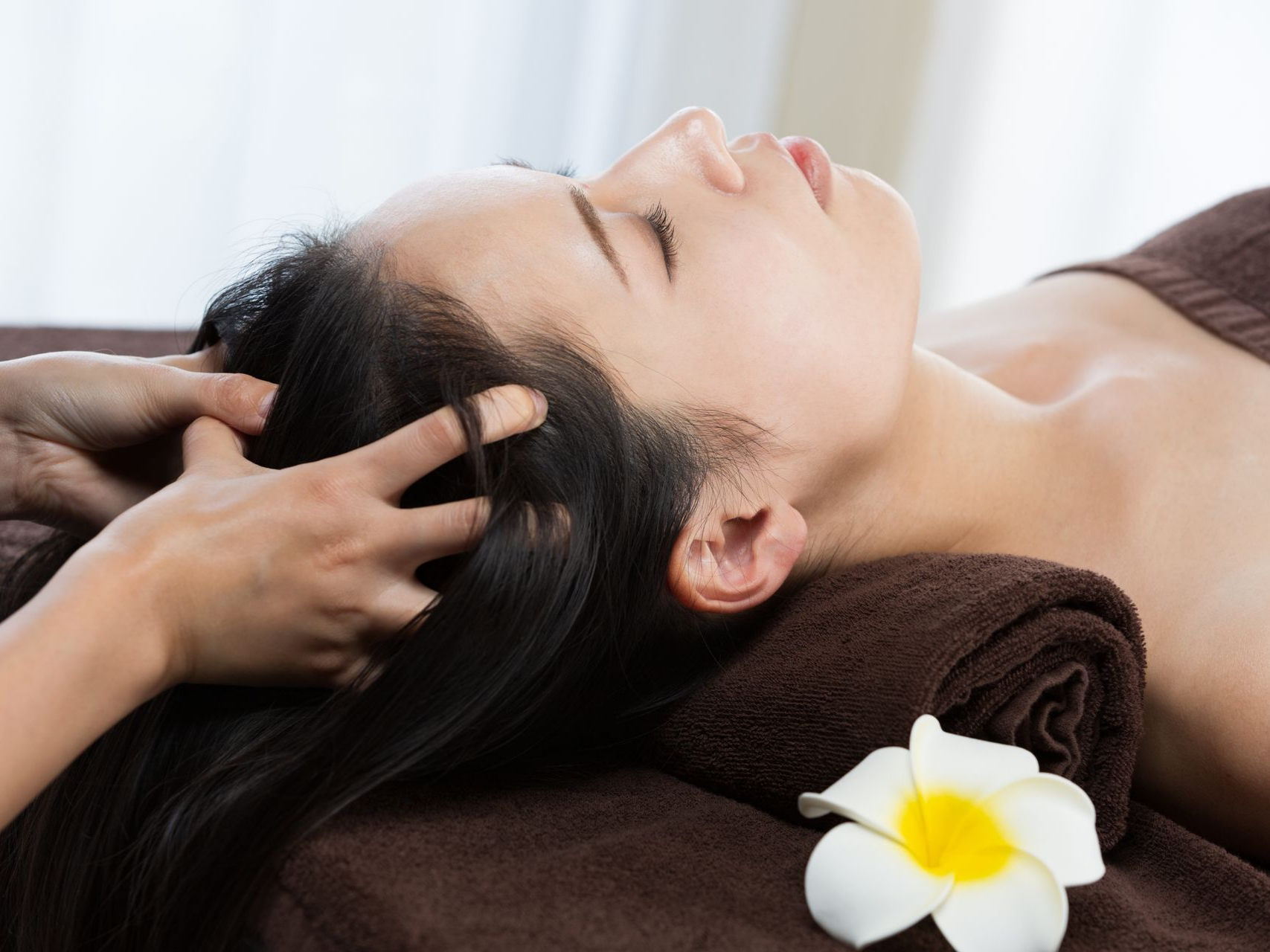 Personne recevant un massage du cuir chevelu ; allongée sur une serviette avec une fleur, les yeux fermés.