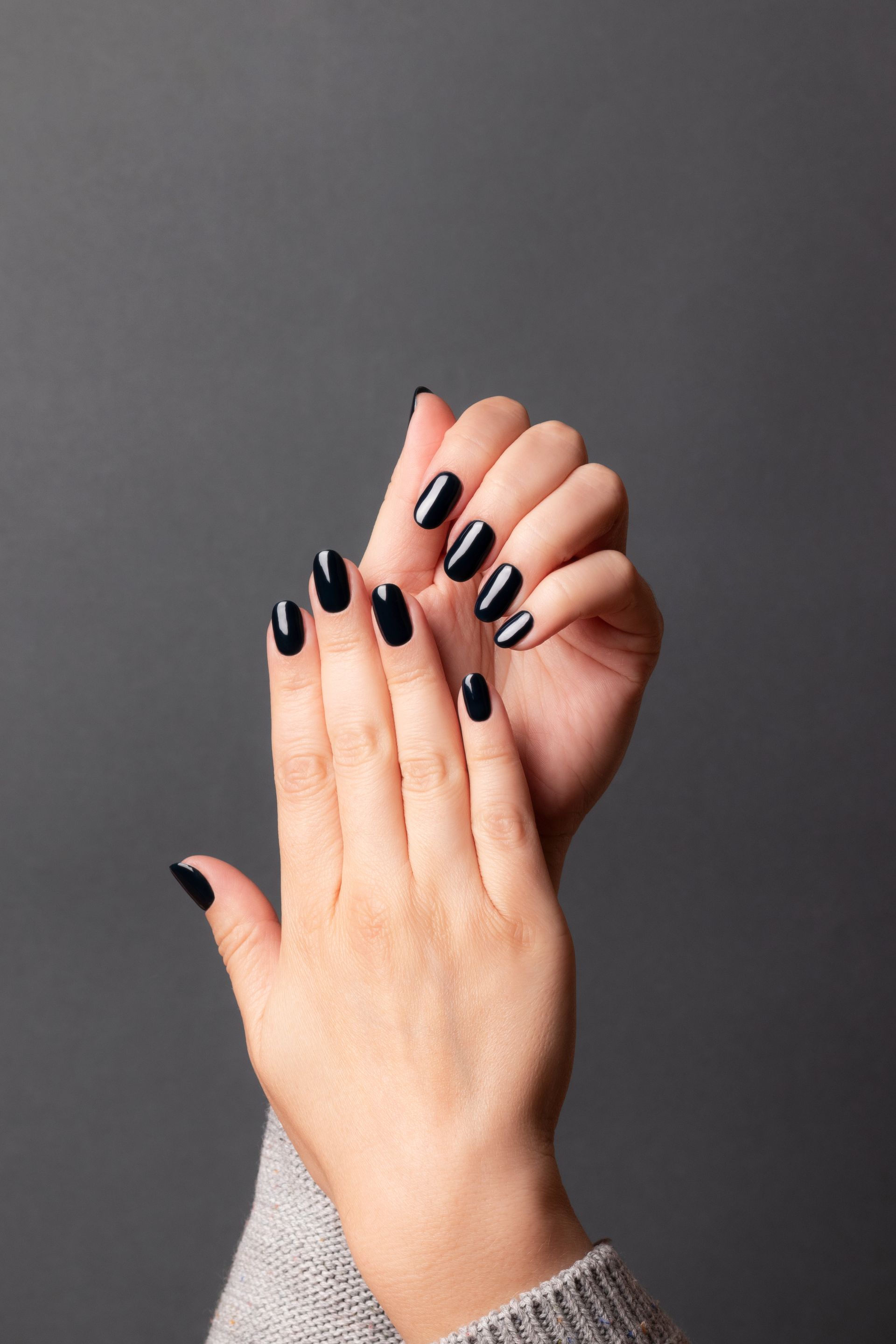 Mains aux ongles foncés, brillants et ovales sur fond gris.