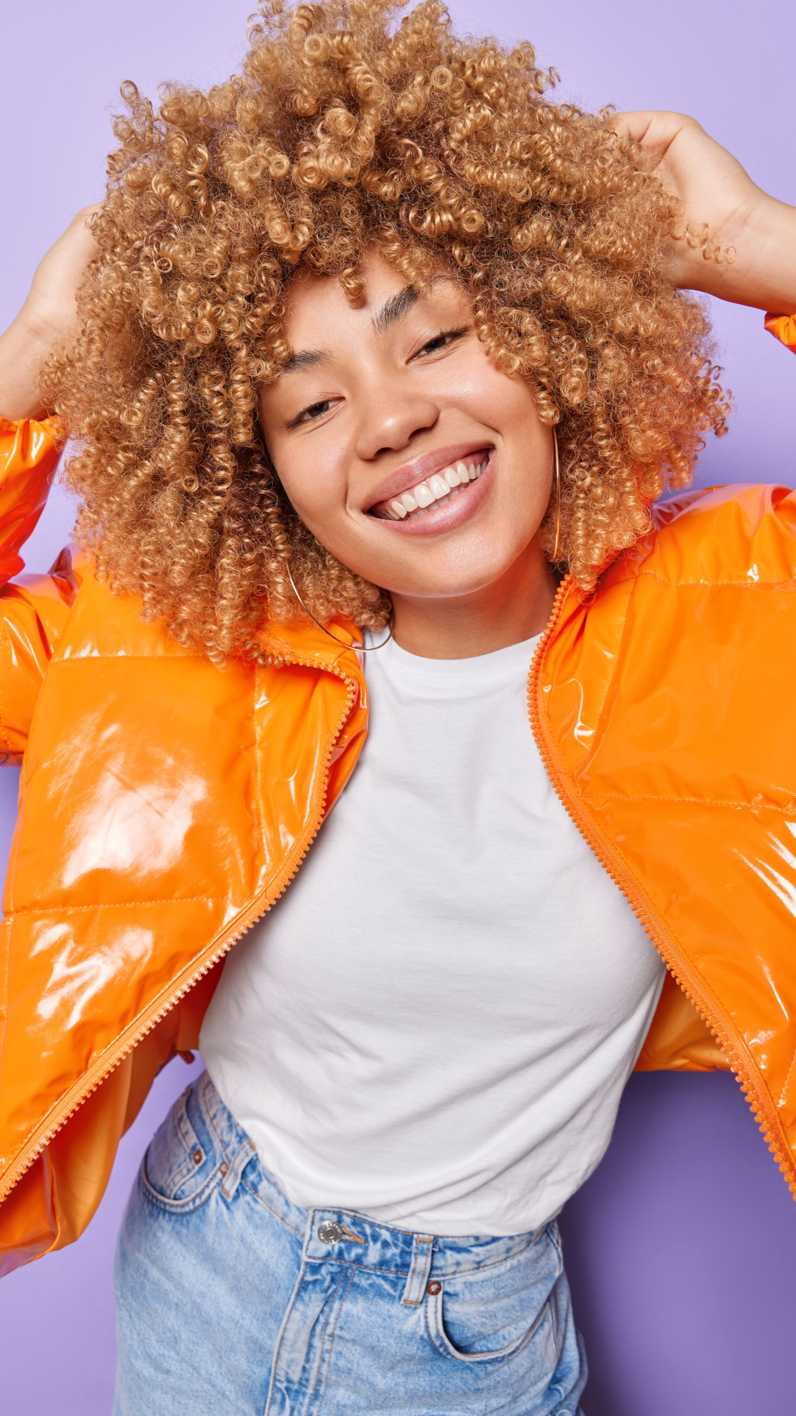 Femme souriante, les mains dans les cheveux bouclés, portant une veste orange et une chemise blanche sur fond violet.