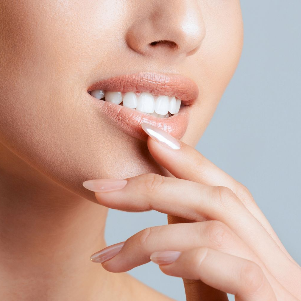 Une femme porte la main à ses lèvres, souriante et dévoilant des dents blanches, sur un fond bleu clair.