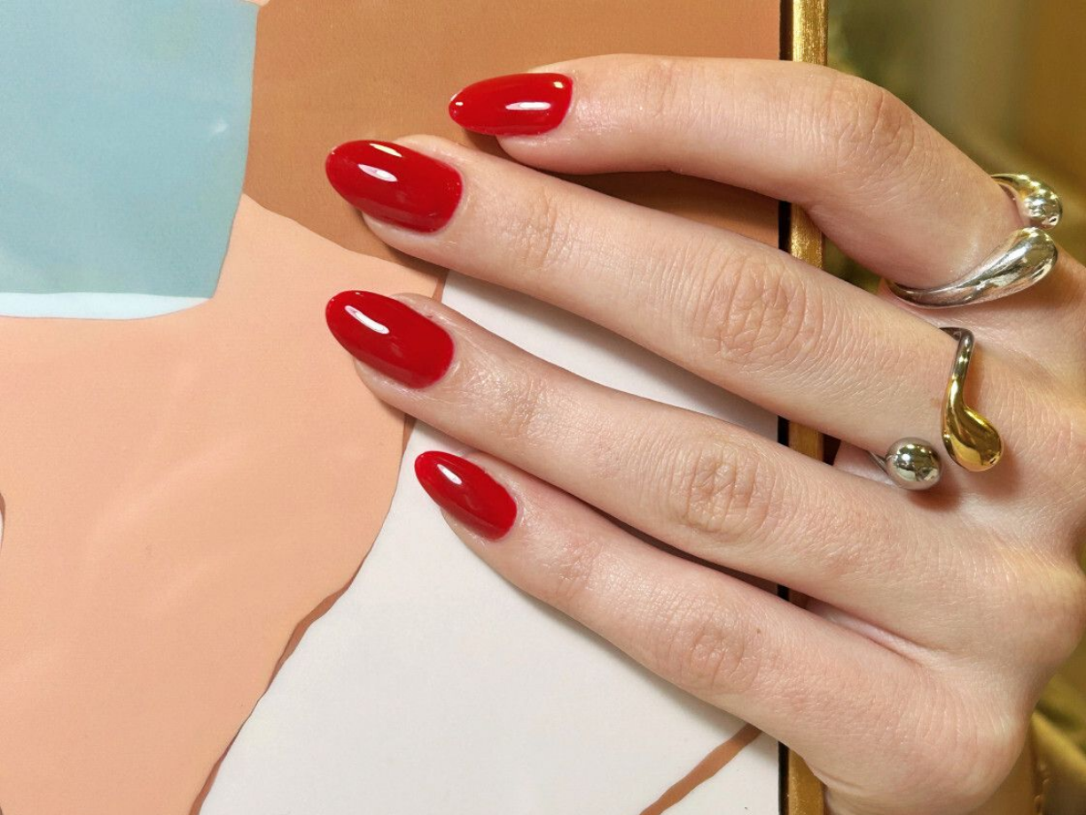 Gros plan sur une main aux ongles ovales, vernis en rouge, portant des bagues en argent et en or, posée sur une surface beige et bleu clair.