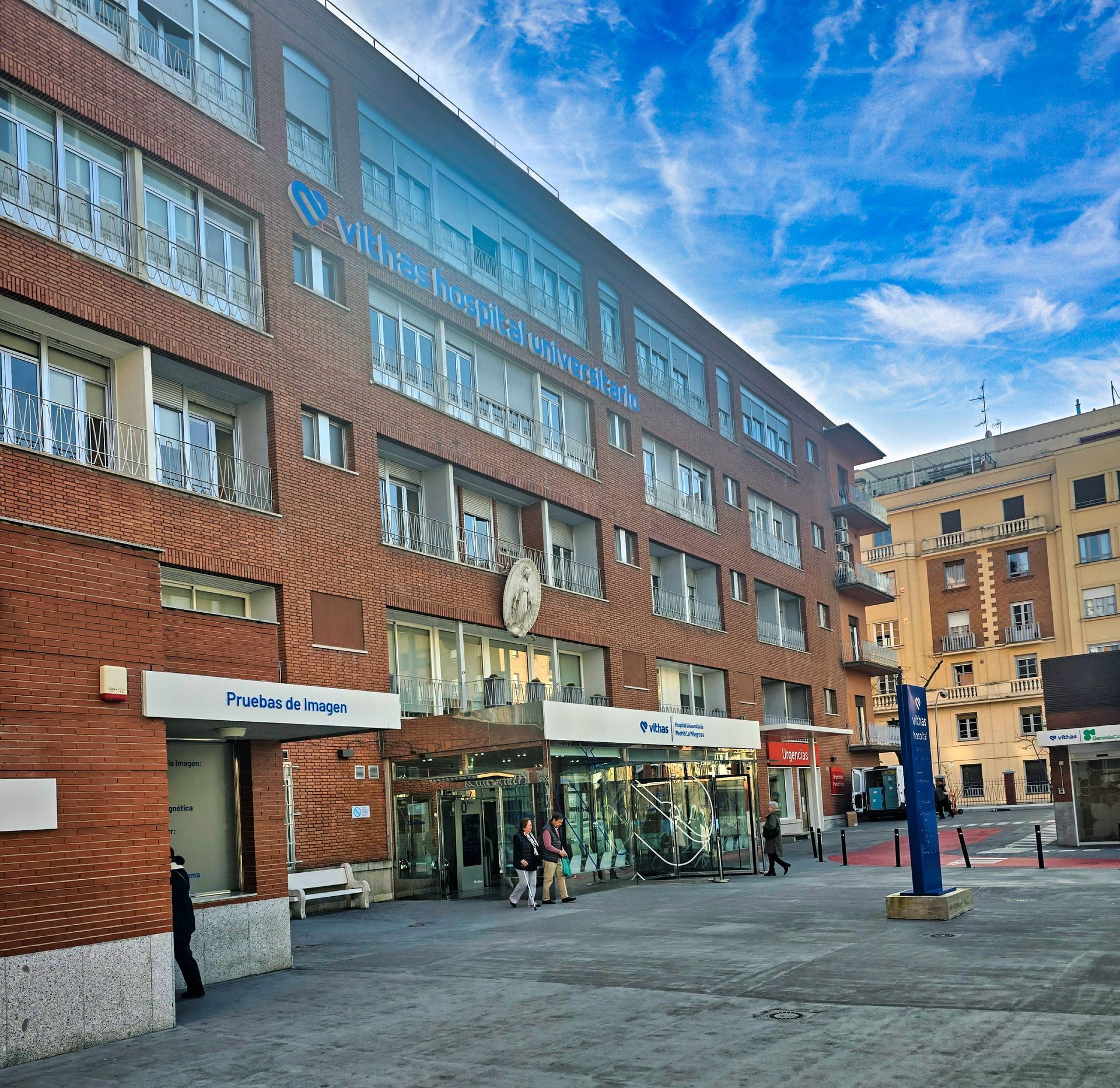 Calle urbana con el edificio del Hospital La Milagrosa de Madrid  y un cielo azul con nubes dispersas.