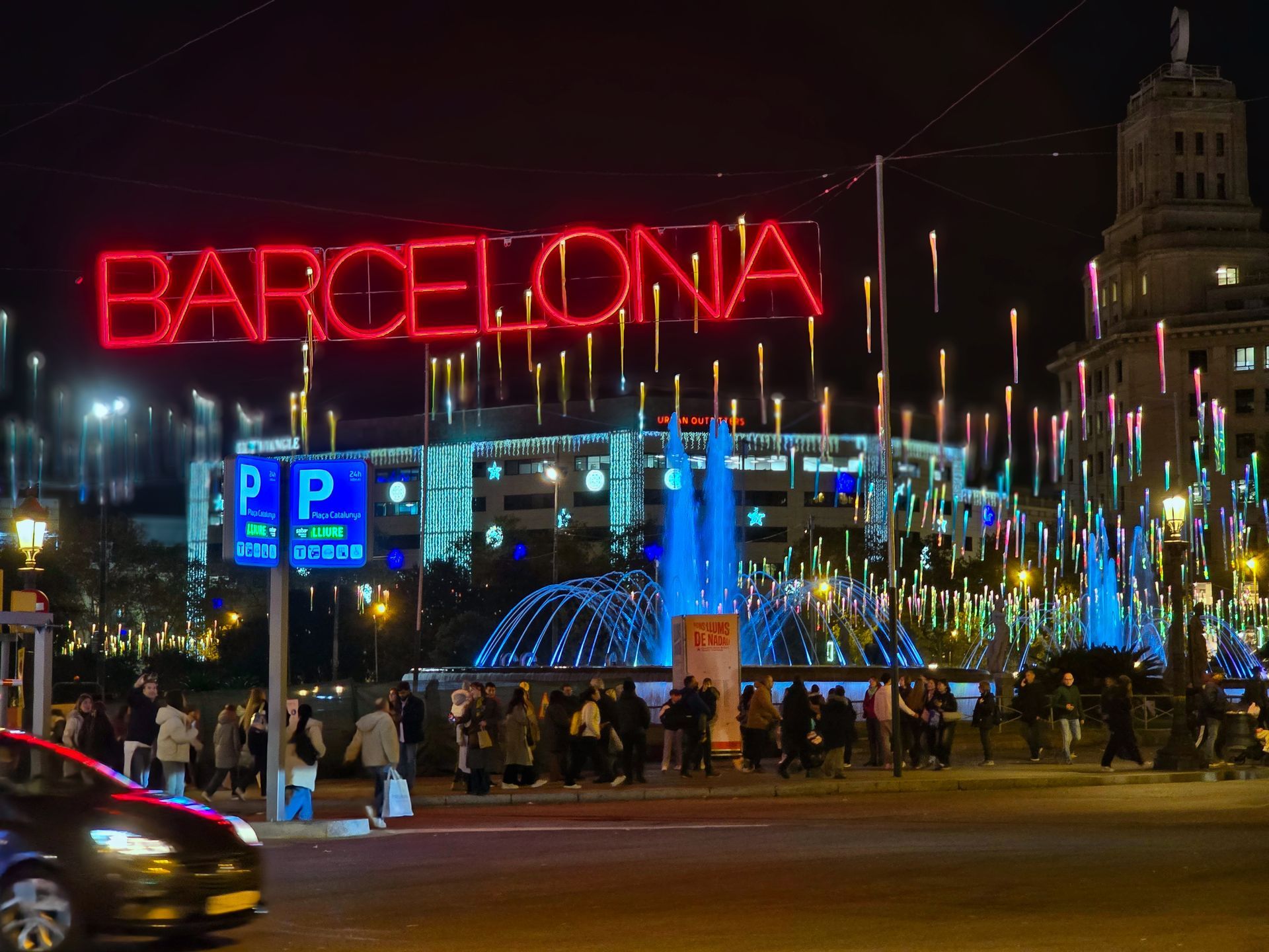 Cartel nocturno de Barcelona que brilla en rojo sobre una plaza concurrida con fuentes y luces de la ciudad.