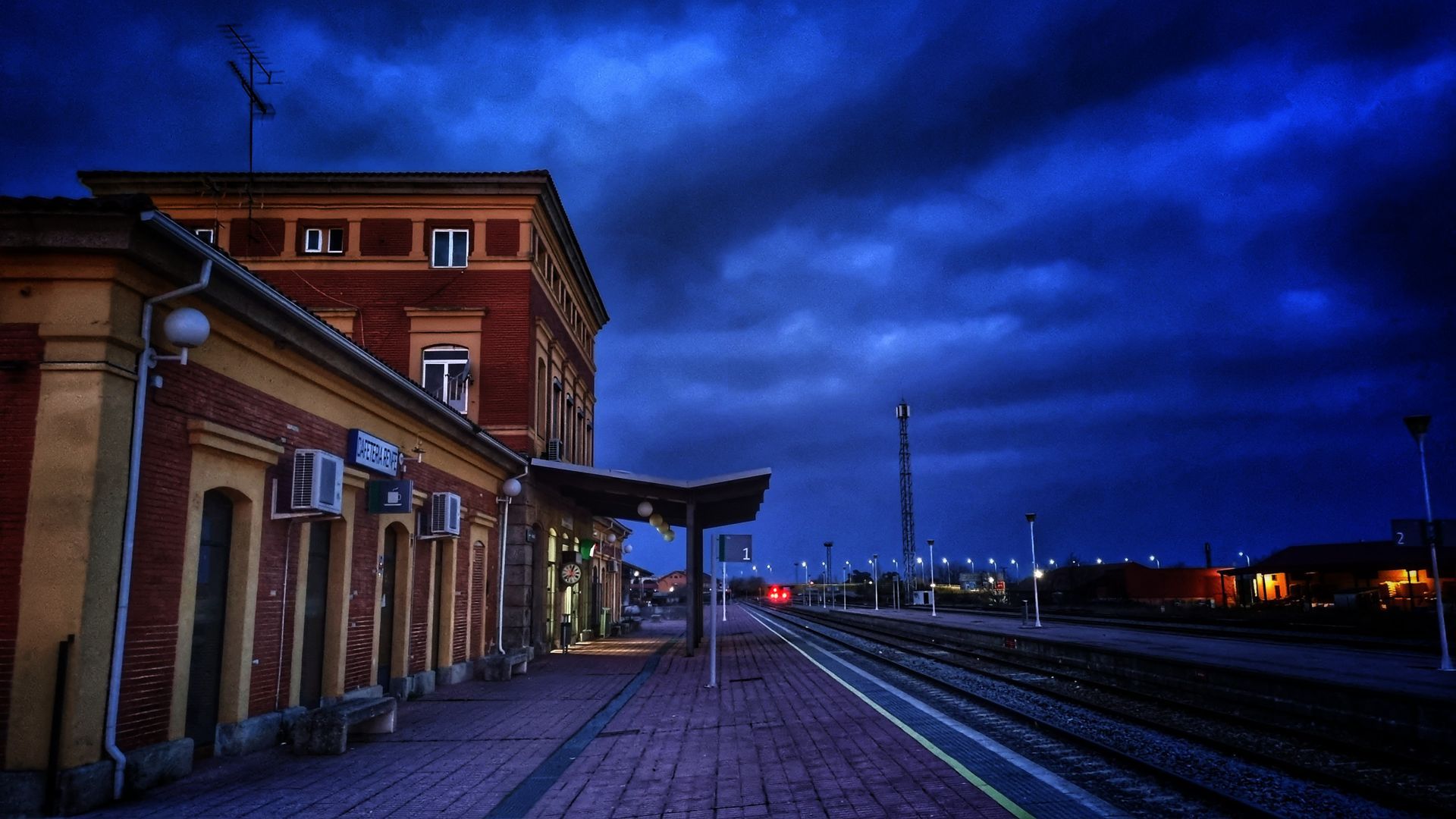 Vista al atardecer de un edificio de estación de Navalmoral de la Mata junto a vías vacías bajo un dramático cielo nublado.
