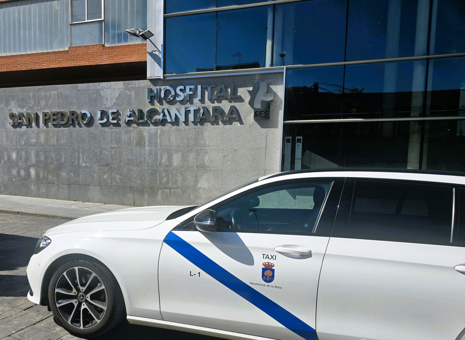 Taxi de Navalmoral blanco con franja azul frente a la puerta del Hospital San Pedro de Cáceres