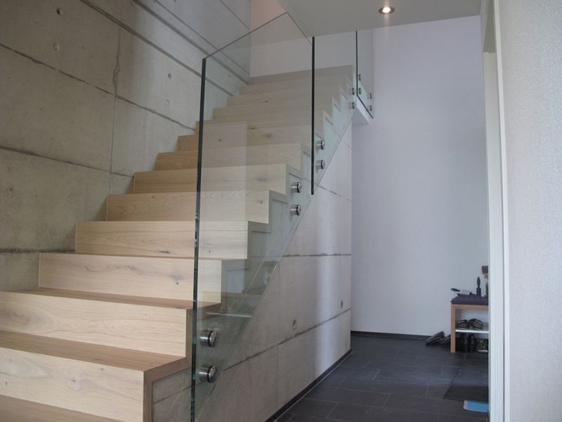 Holztreppe mit Glasgeländer vor einer Beton- und weißen Wand.