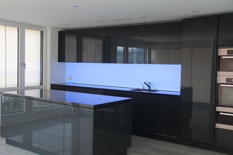 Moderne Küche mit schwarzen Hochglanzschränken, Kochinsel & blau beleuchtetem Fliesenspiegel. Fenster links, Backöfen rechts.