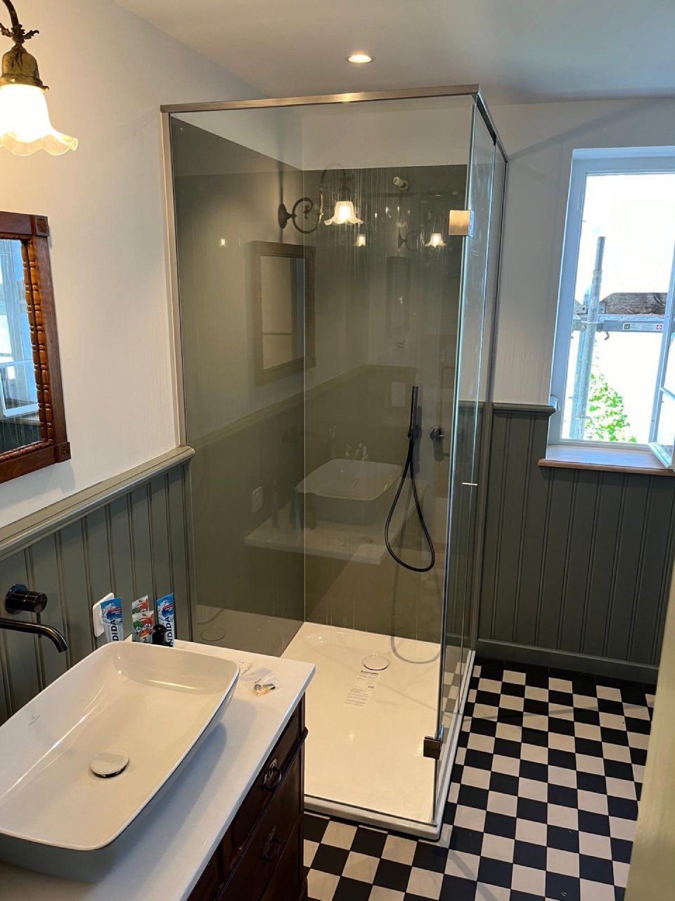 Badezimmer mit Glasdusche, weißem Waschbecken, Schachbrettboden und dunkelgrüner Wandvertäfelung.