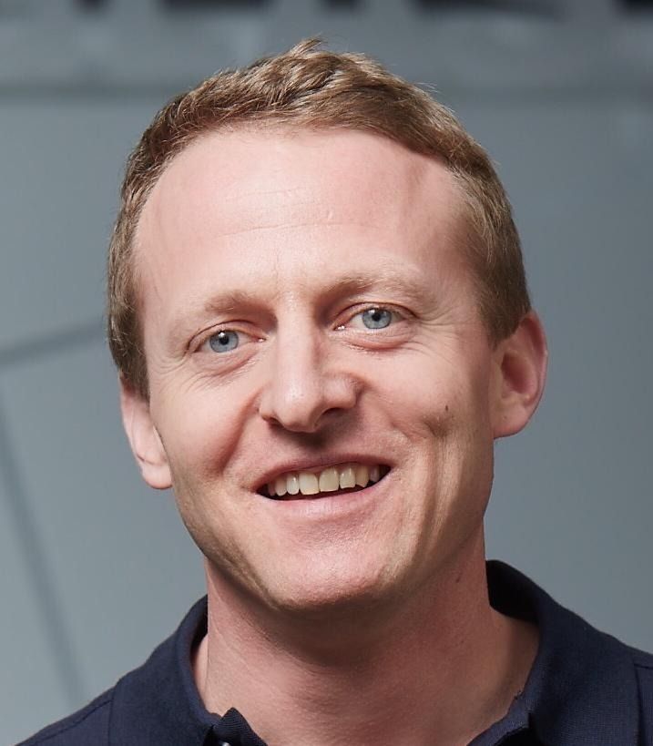 Ein Mann mit heller Haut lächelt in die Kamera; er trägt ein dunkelblaues Hemd. Die Szene spielt sich in einem Innenraum ab.