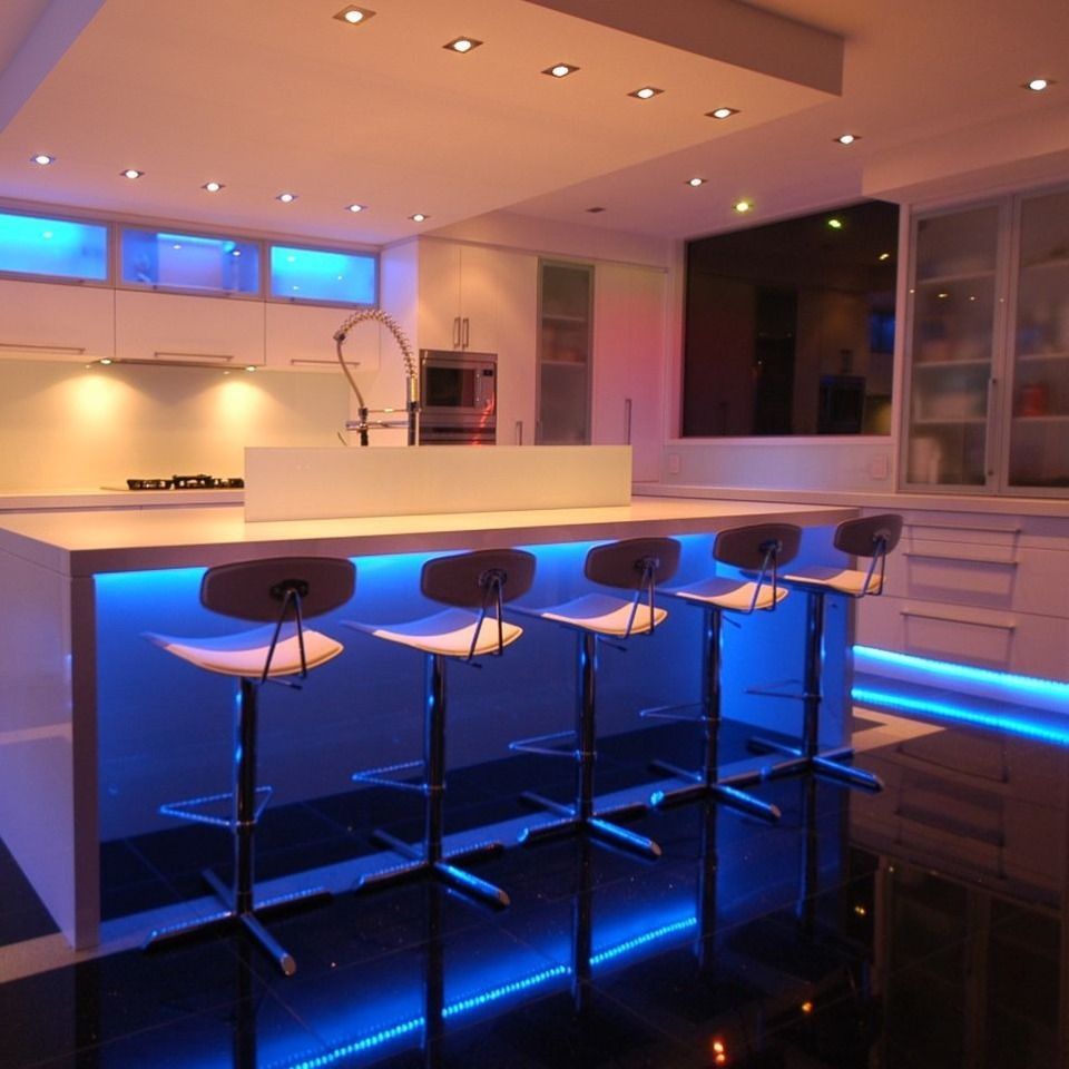 Moderne Küche mit weissen Schränken, Kochinsel und Barhockern; beleuchtet mit blauen und orangen LED-Leuchten.
