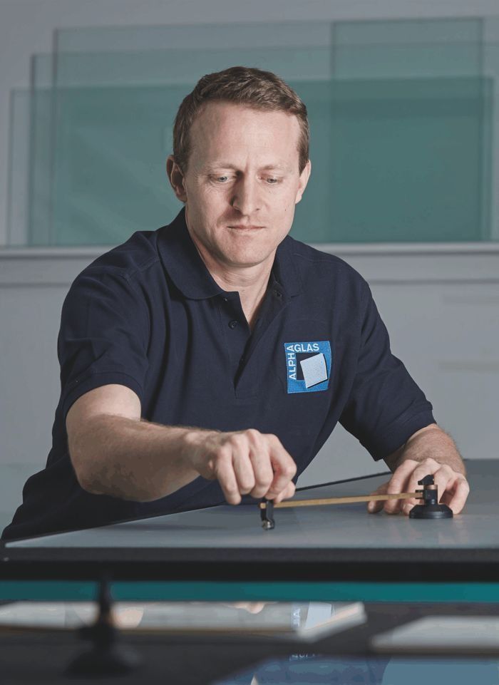 Ein Mann mit heller Haut lächelt in die Kamera; er trägt ein dunkelblaues Hemd. Die Szene spielt sich in einem Innenraum ab.