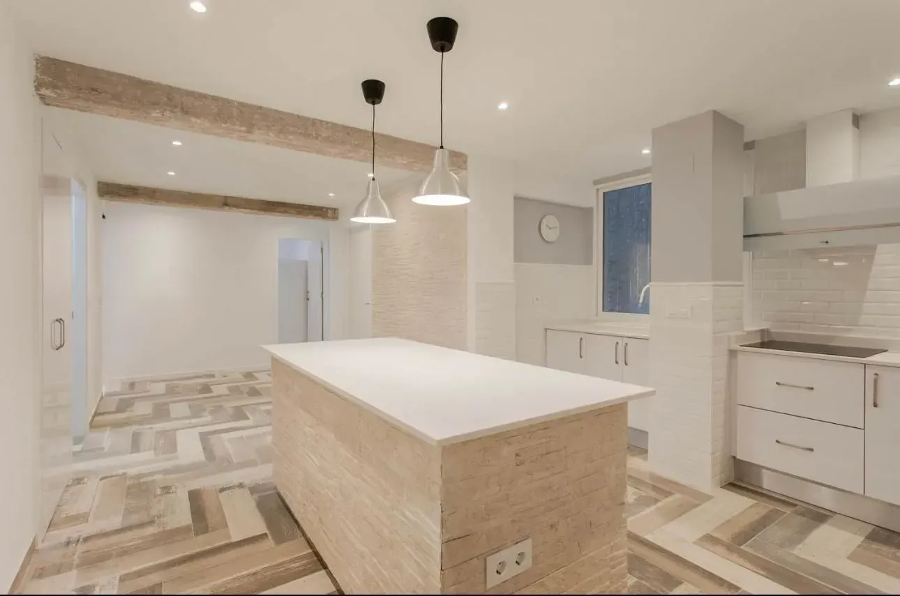 Cocina abierta con suelo de madera, armarios blancos e isla. Dos lámparas colgantes cuelgan sobre la isla.