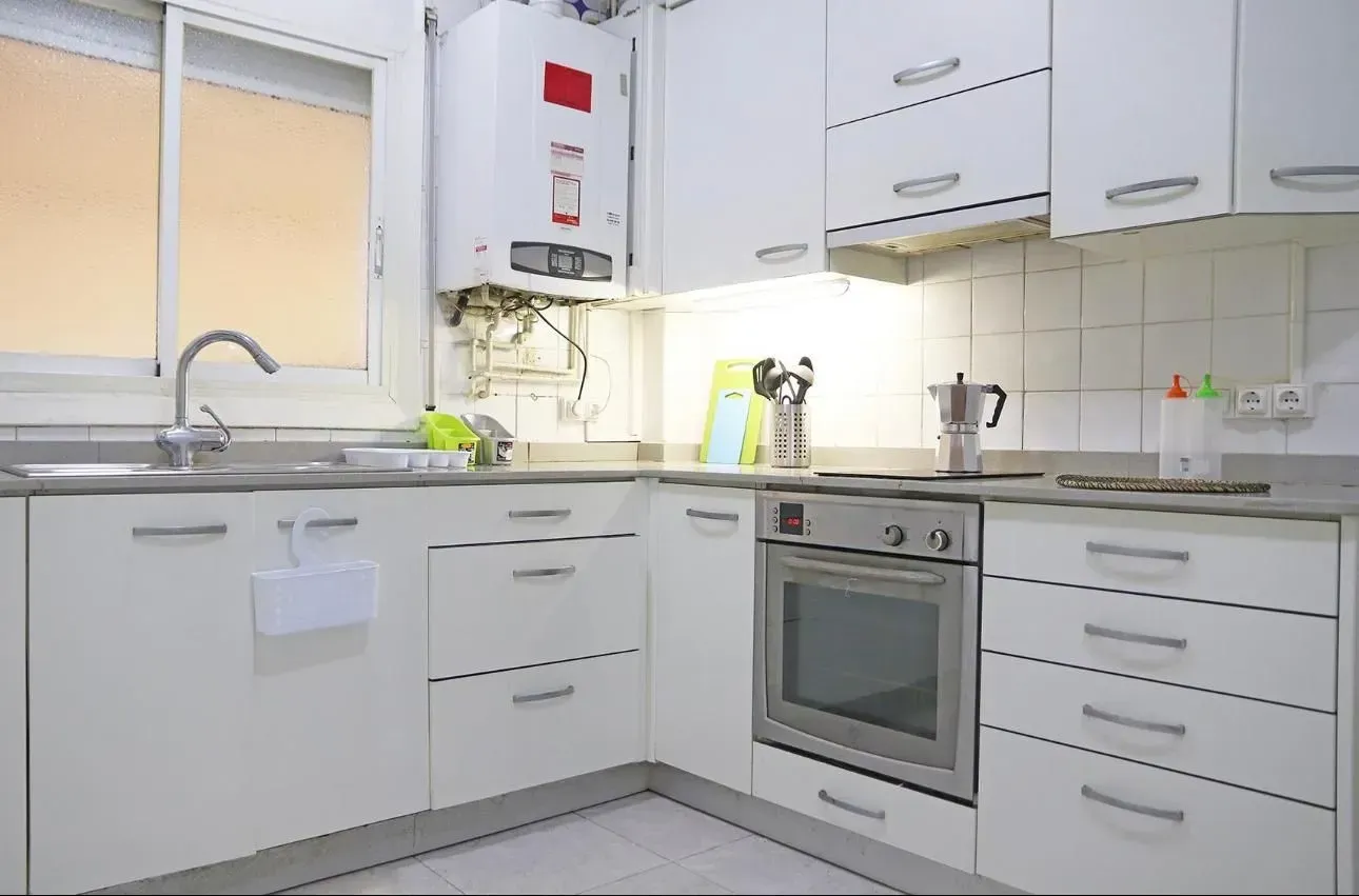 Cocina blanca con armarios, fregadero y horno. Calentador de gas montado en la pared.