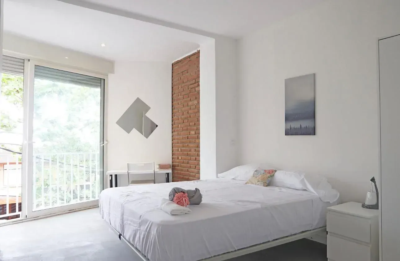 Dormitorio con cama grande, balcón, pared de ladrillo visto, paredes blancas y decoración minimalista.
