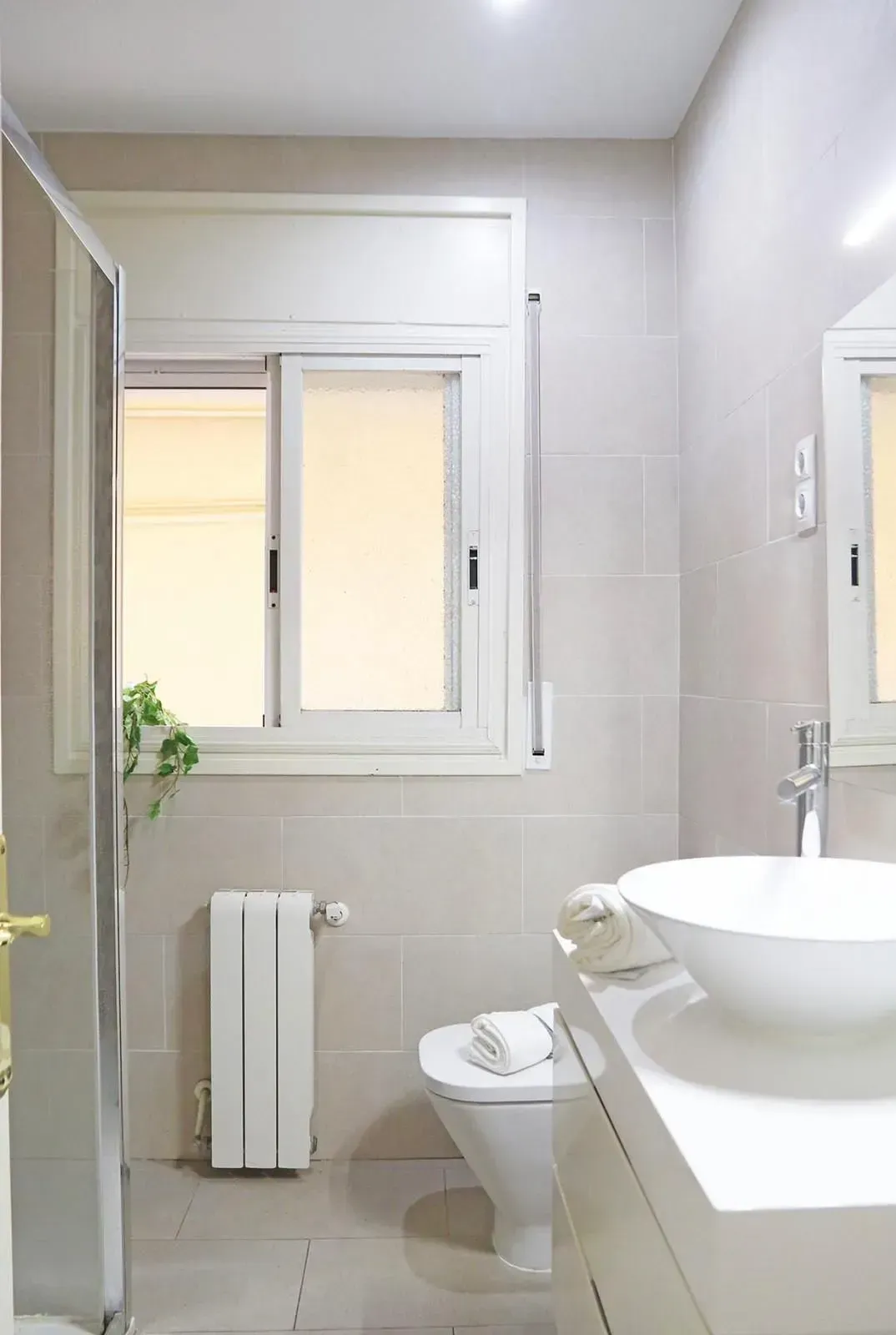 Baño con inodoro, lavabo y ventana blancos. Se ven paredes de azulejos beige, un radiador y una mampara de ducha de cristal.
