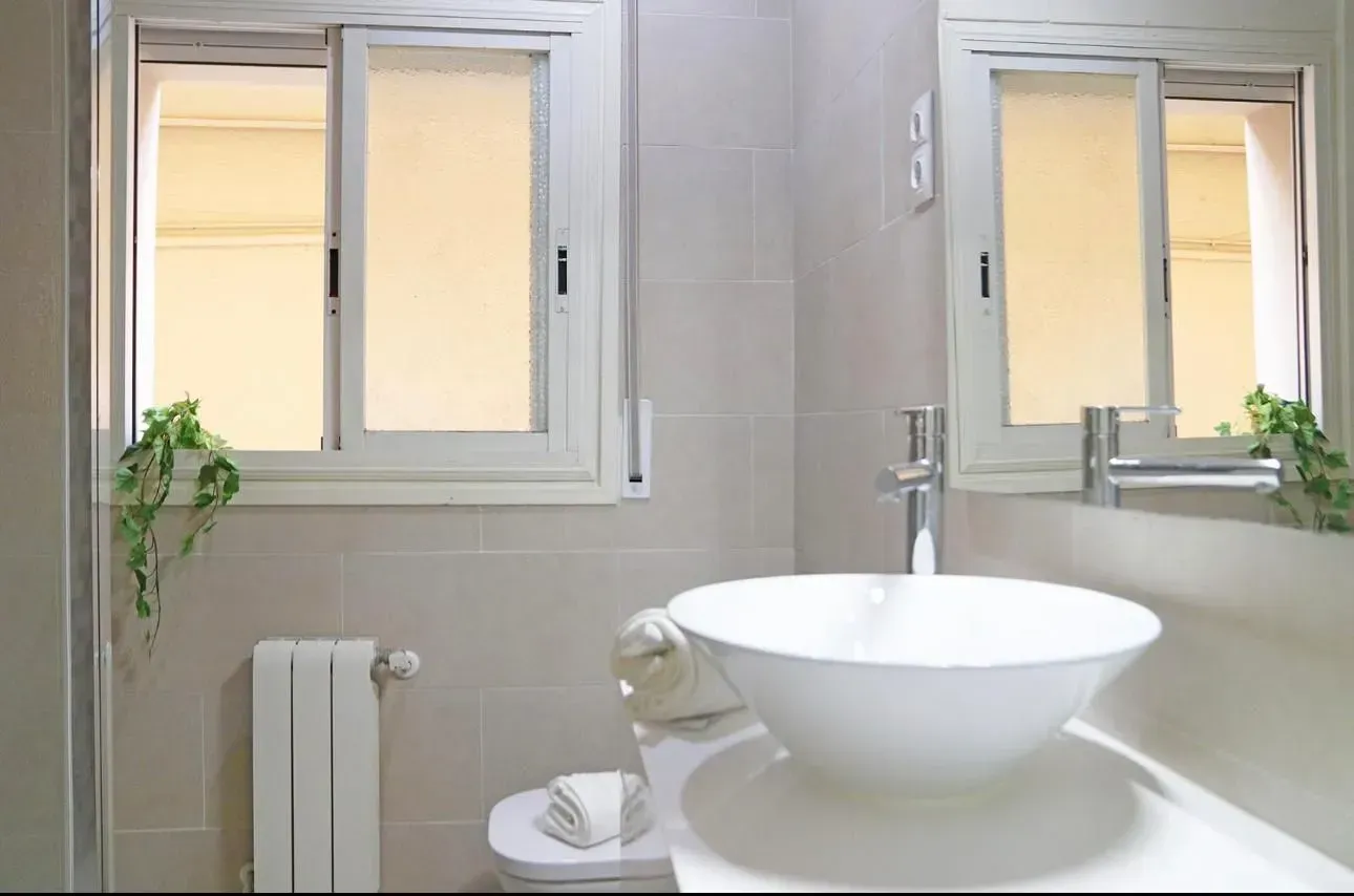 Baño con lavabo blanco, espejo, dos ventanas y azulejos gris claro.