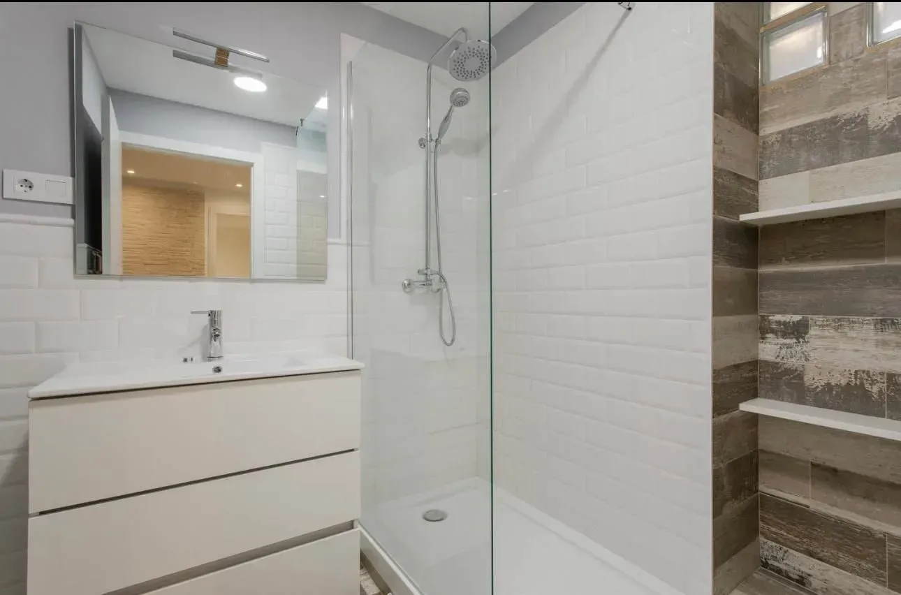 Baño moderno con azulejos blancos y grises, ducha de cristal, tocador y espejo.