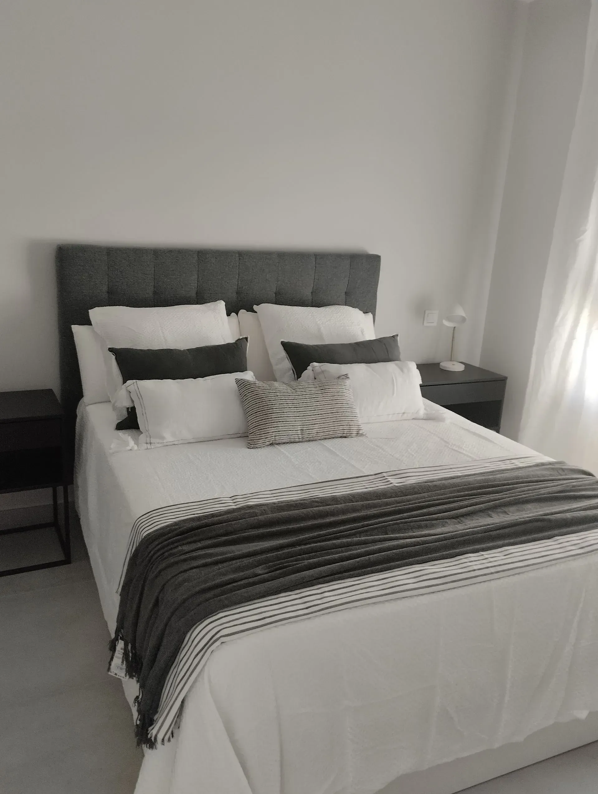Dormitorio con cabecera capitoné gris, ropa de cama blanca, manta oscura y almohadones decorativos.