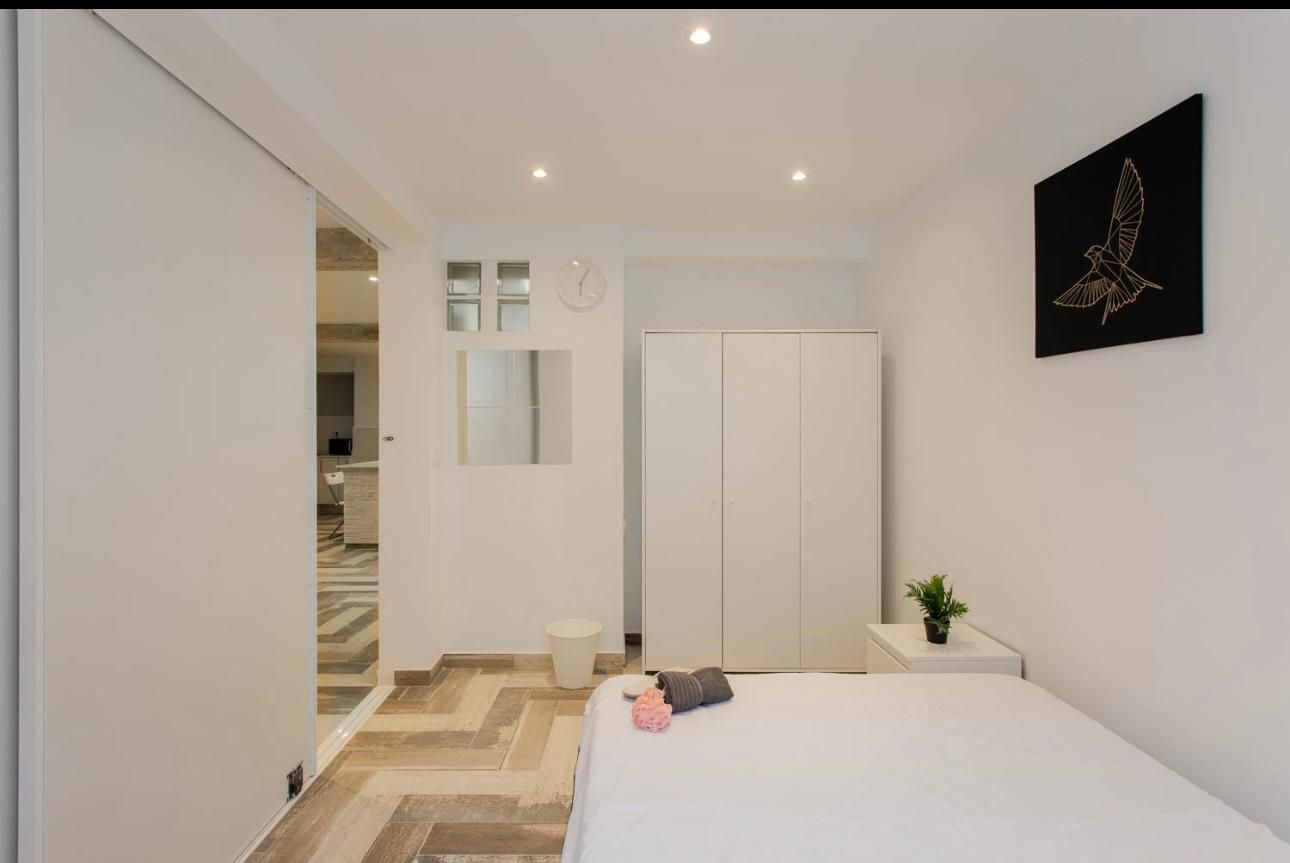 Dormitorio blanco con suelo en espiga, cama, armario y obras de arte.