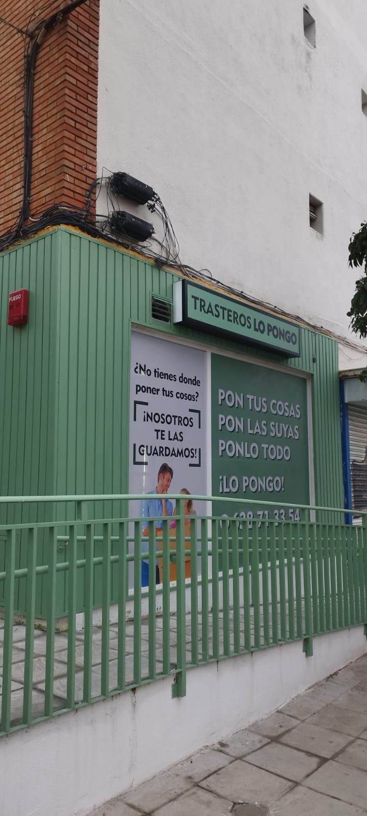 Edificio ecológico con letreros y valla metálica. Pared de ladrillo y edificio blanco al fondo.