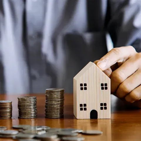 Una persona sostiene una casa de madera junto a pilas de monedas sobre una mesa.