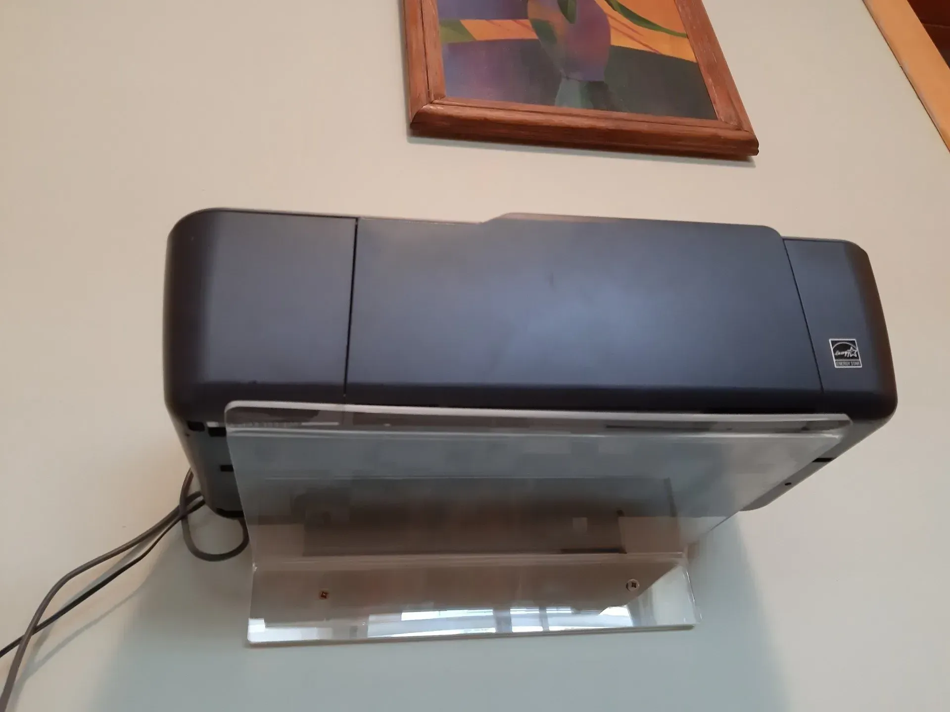 Una impresora negra está colocada sobre un soporte transparente en una pared.