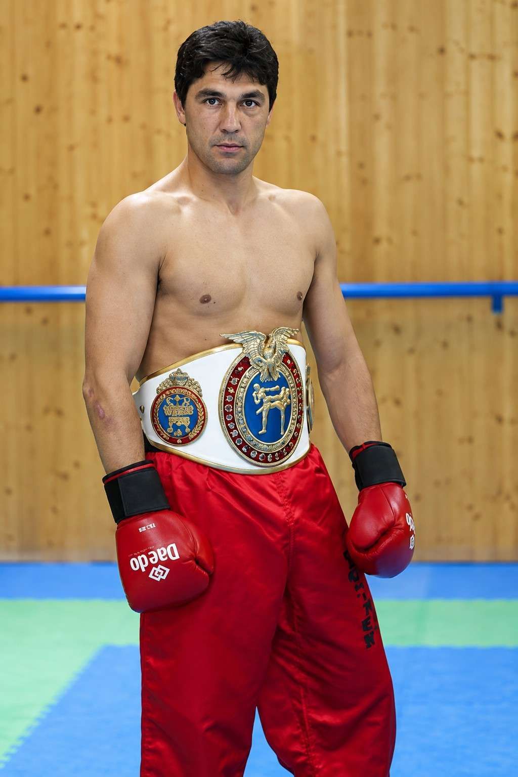 Un boxeador con guantes rojos y pantalones cortos rojos, luciendo cinturones de campeón, está de pie en un gimnasio.
