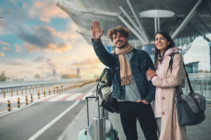 Couple à l'aéroport faisant signe de la main ; homme avec sac, femme avec sac à main ; bagages à proximité, soleil couchant.