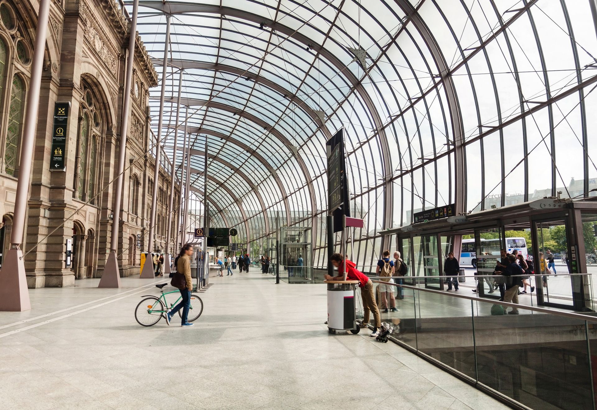 Un hall de gare couvert avec un plafond de verre voûté. Les piétons, les cyclistes et les piétons attendent près de l'entrée.