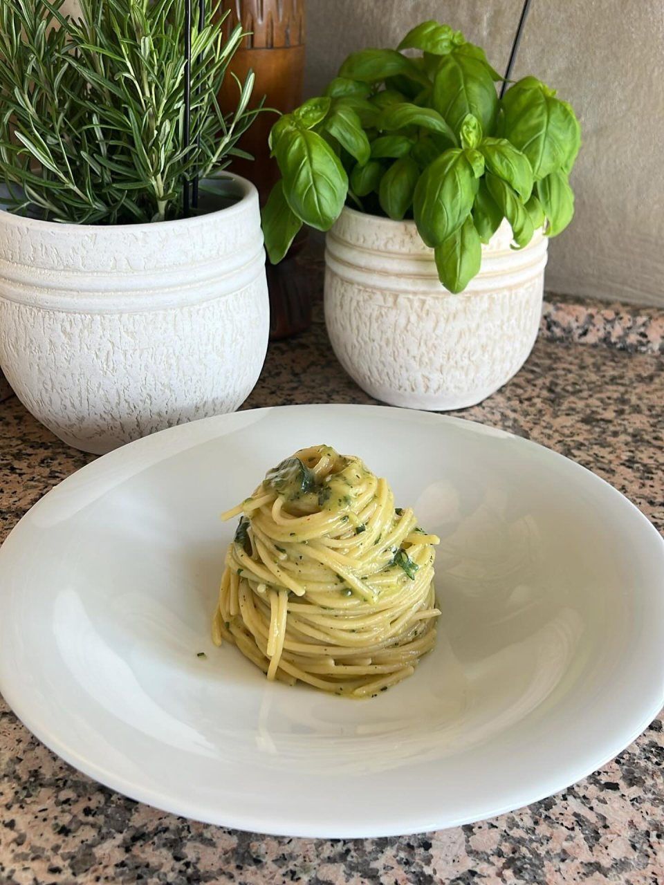 Pasta mit Pesto auf einem weissen Teller, im Hintergrund Rosmarin und Basilikum im Topf.