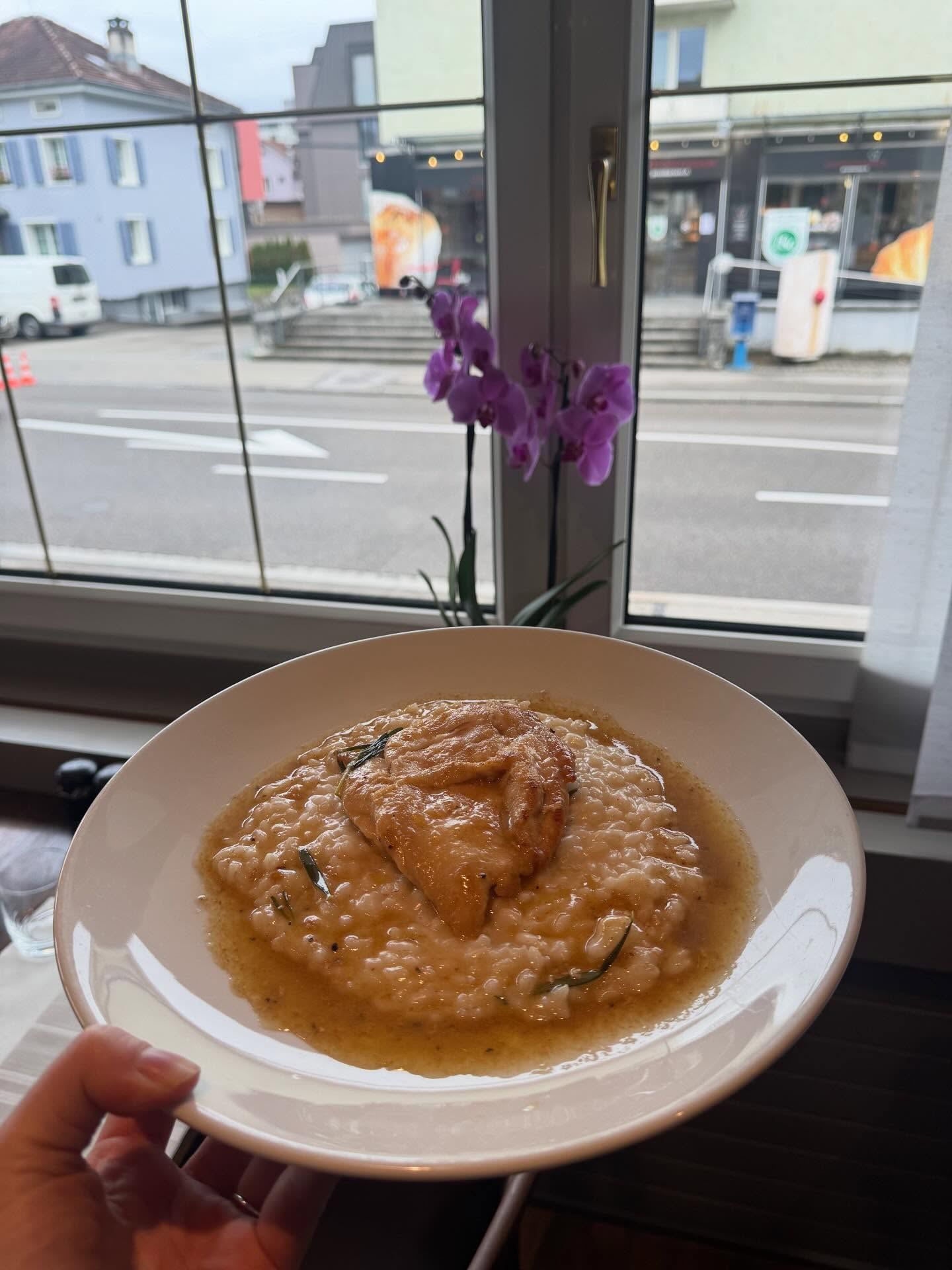 Ein Teller Risotto mit Topping wird vor einem Fenster mit Blick auf eine Strasse mit Gebäuden gehalten.