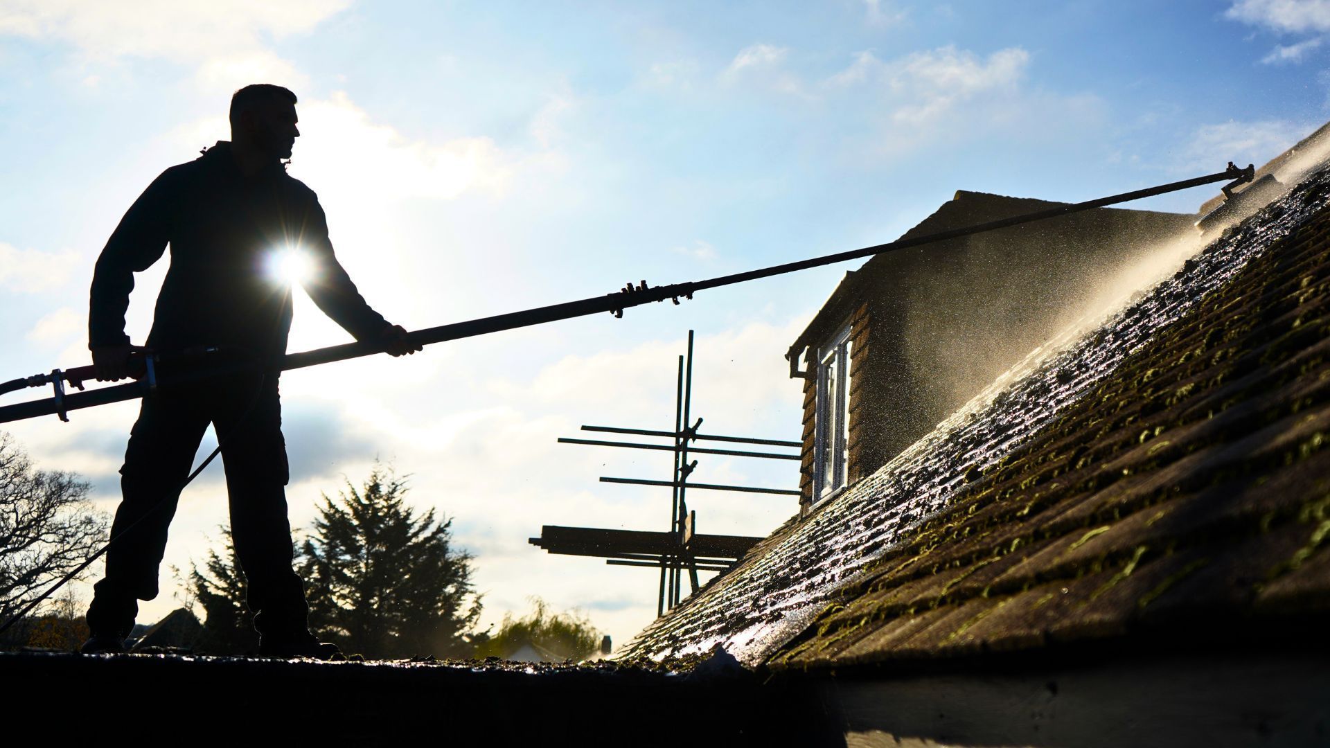 Worker power-washing a roof at sunset, spraying water from a hose beside a house.