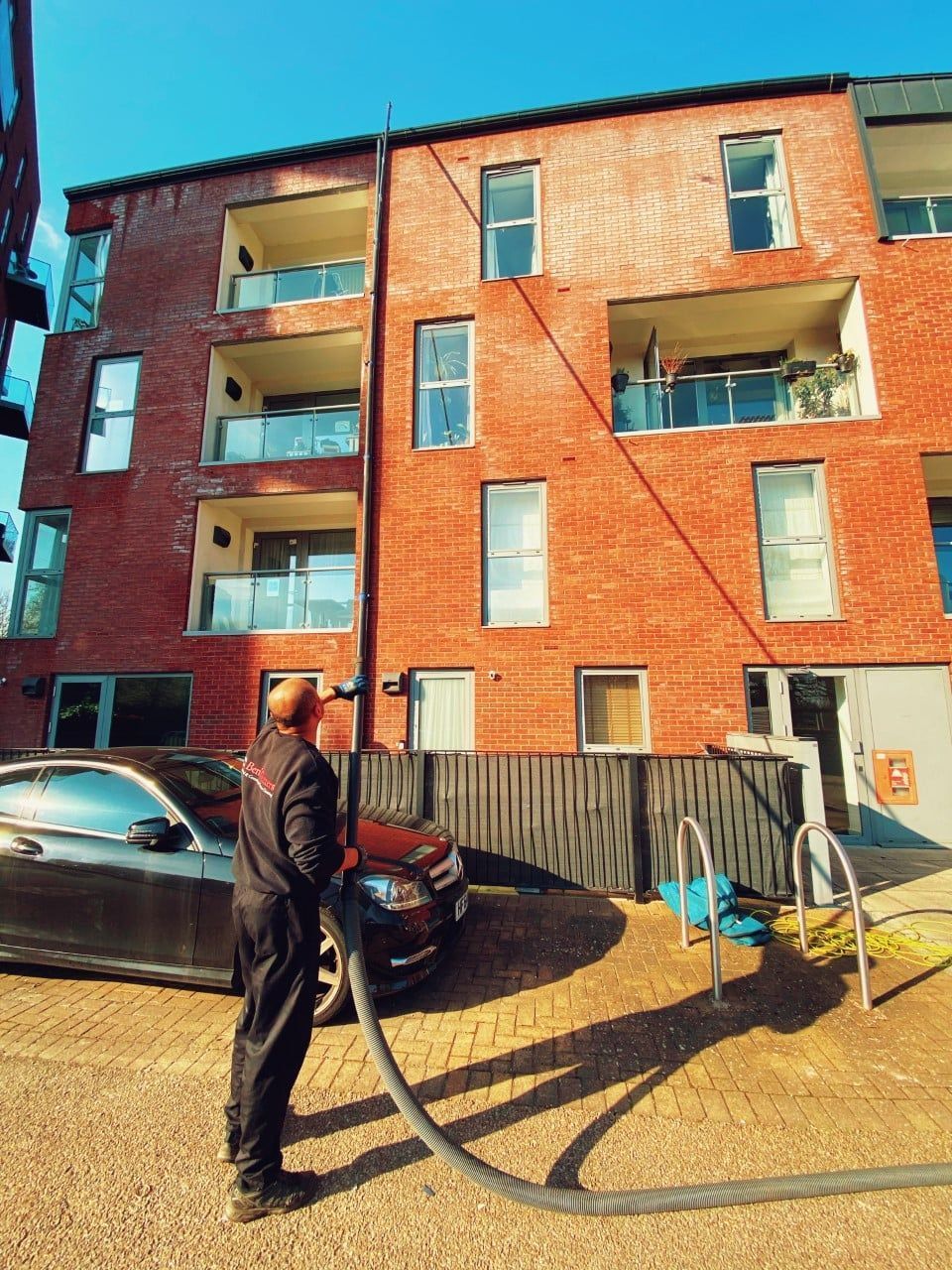 Person standing beside a parked car in front of a red brick apartment building, with balconies and a garden hose.