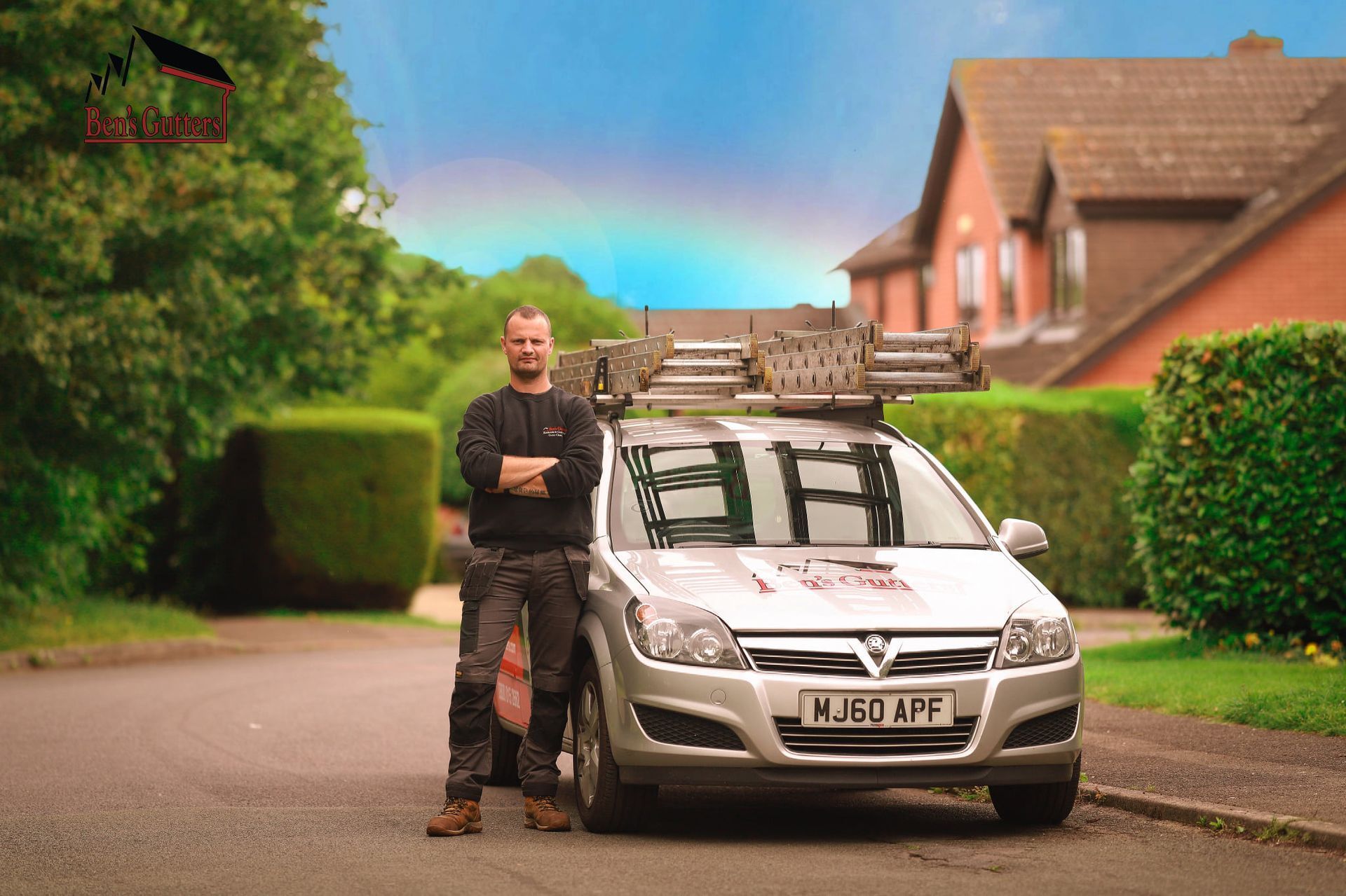 Man standing beside a white car with roof rack in a suburban street, under a rainbow.